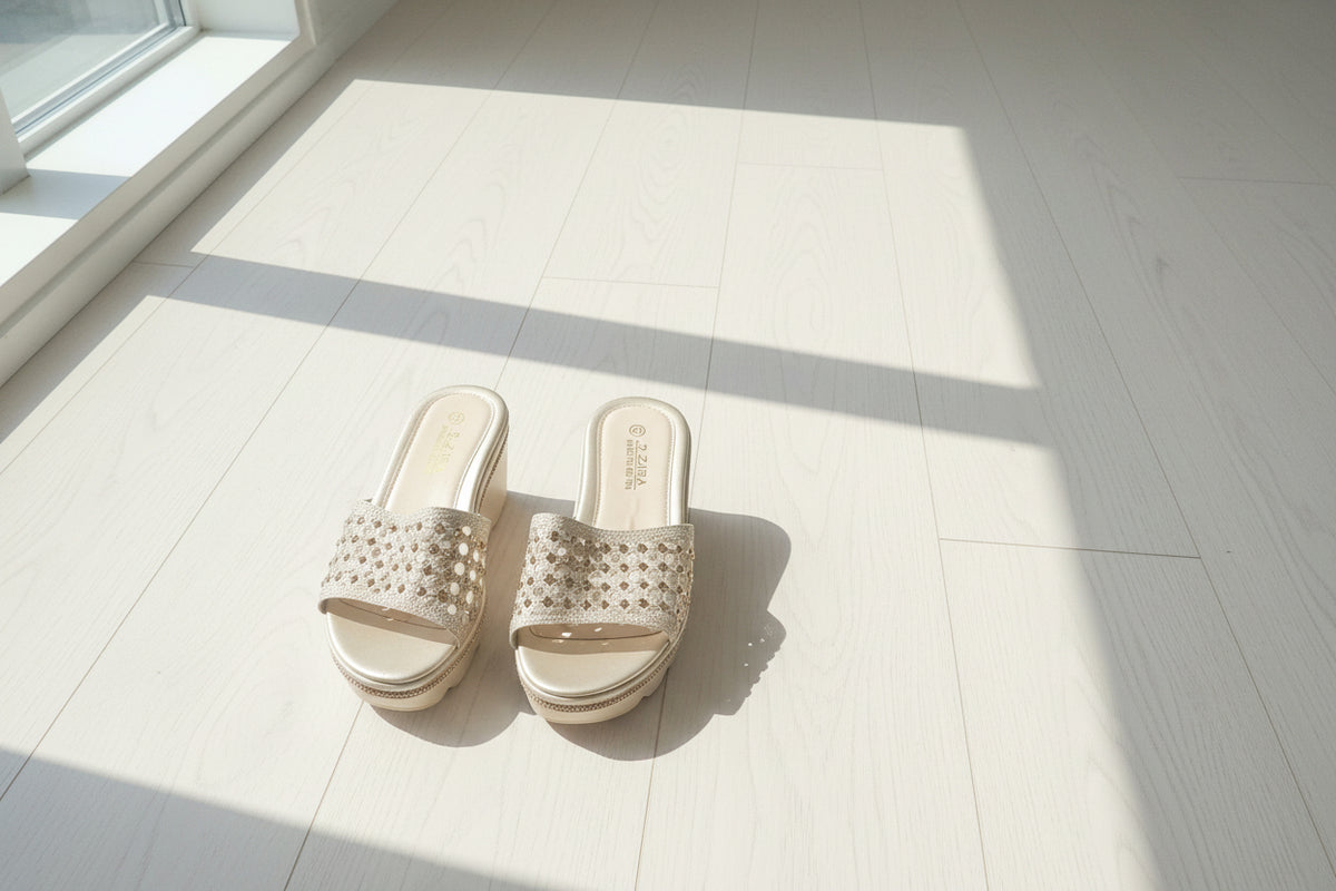 A pair of women's slide sandals with woven beige straps and a low block heel, shown from above on a dark wood plank floor.