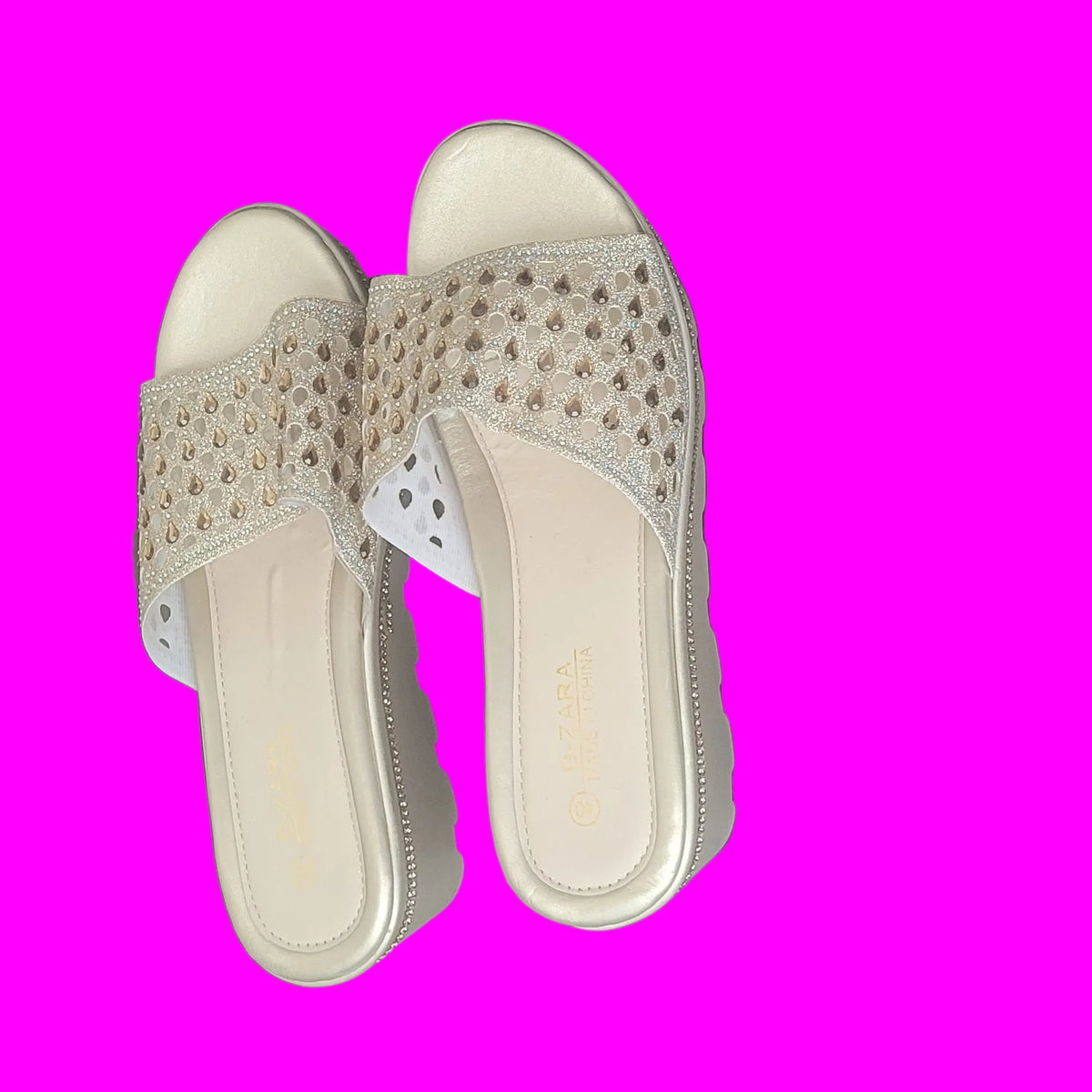 A pair of beige slide sandals with metallic gold star-patterned straps, shown on a dark wood plank floor.