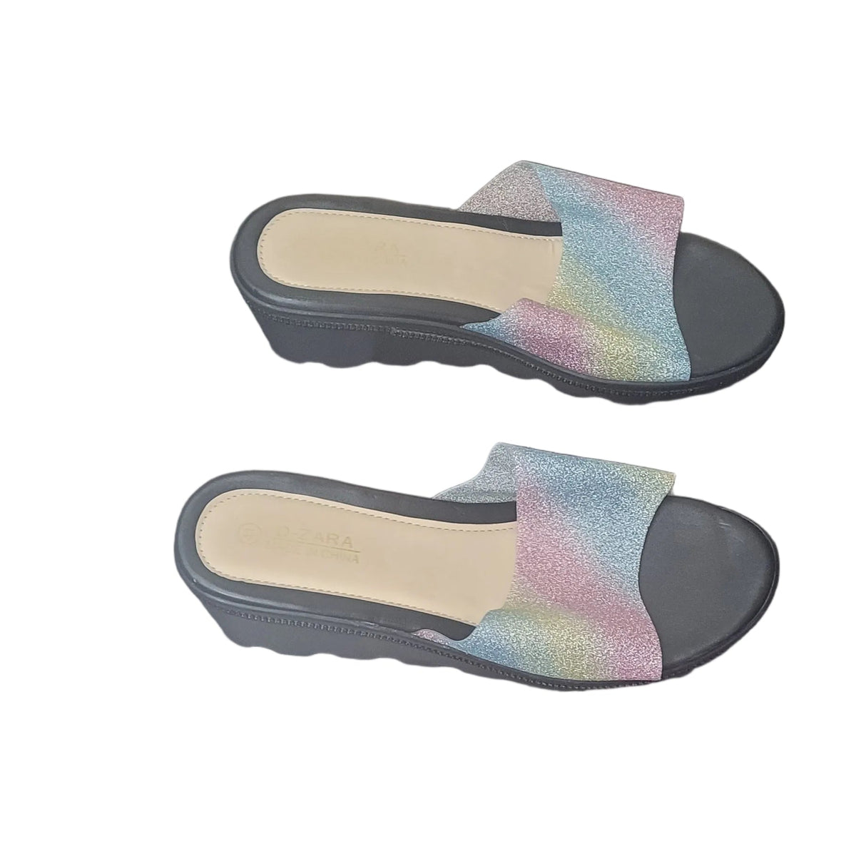 A pair of women's slide sandals with black wedge heels and iridescent, rainbow-colored straps, shown on a dark wood floor.