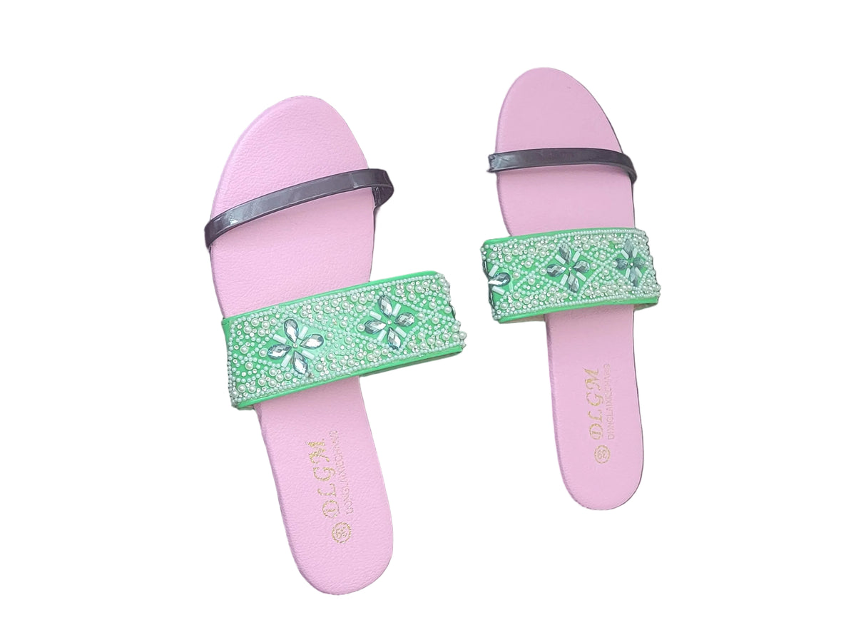 A pair of pink slide sandals with wide green embellished straps, photographed on a dark wood floor.