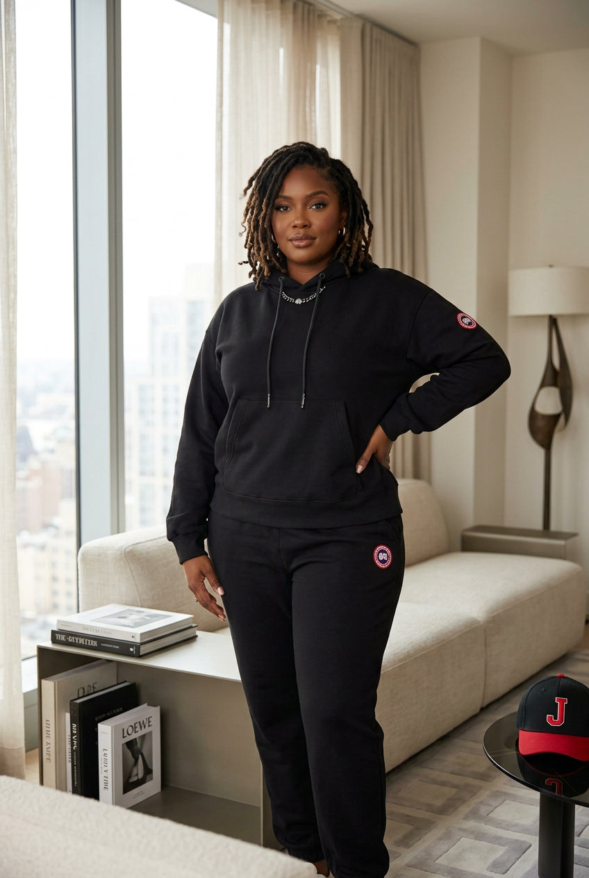 A black two-piece tracksuit set consisting of a pullover hoodie and matching sweatpants, styled on a dark round table with decorative props including a white hat, a magazine, and a silver heart-shaped pendant.
