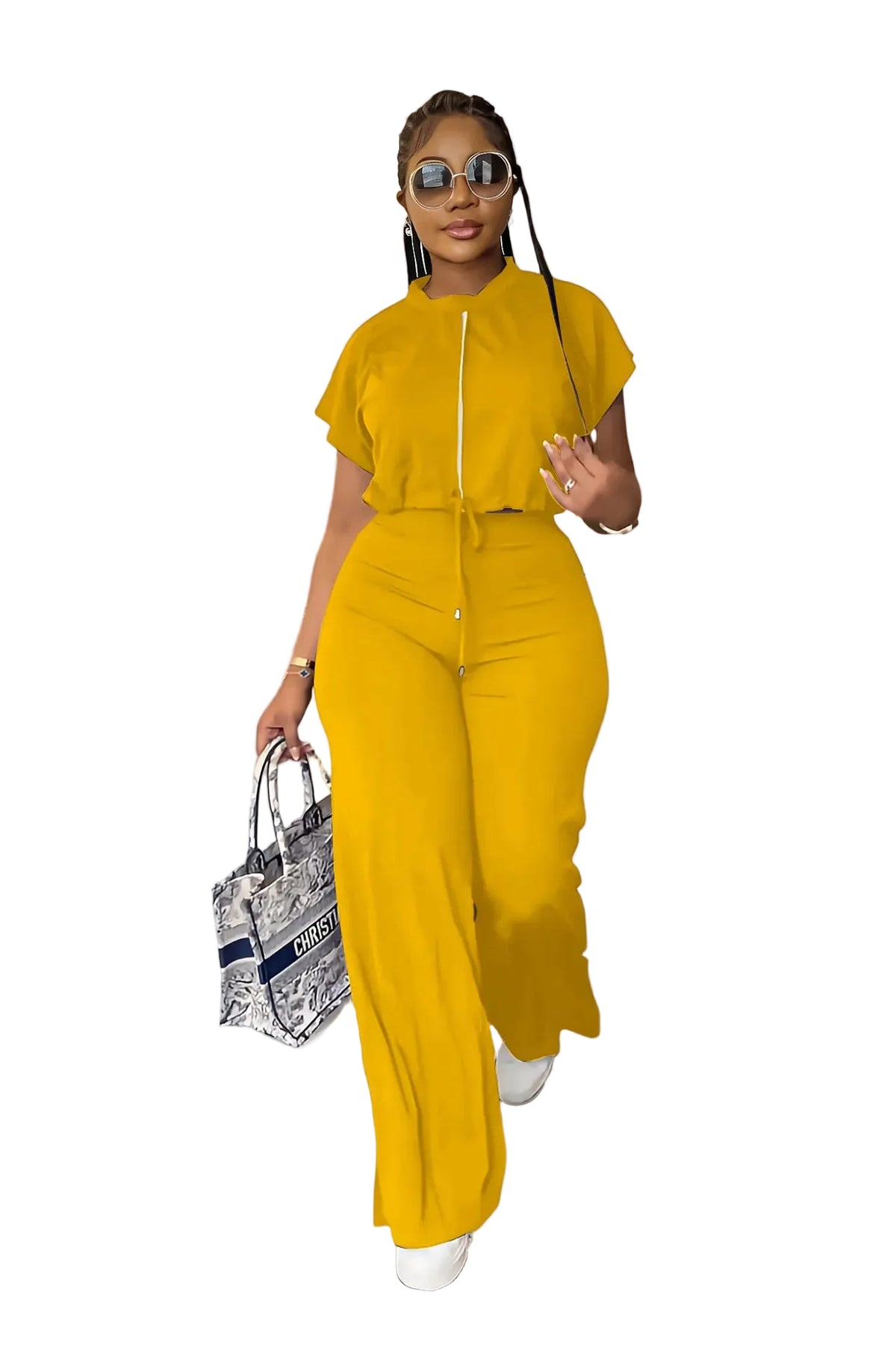 A woman modeling a bright yellow two-piece jumpsuit with a front zipper and tie waist, styled with sunglasses, a patterned handbag, and white sneakers in an indoor setting.