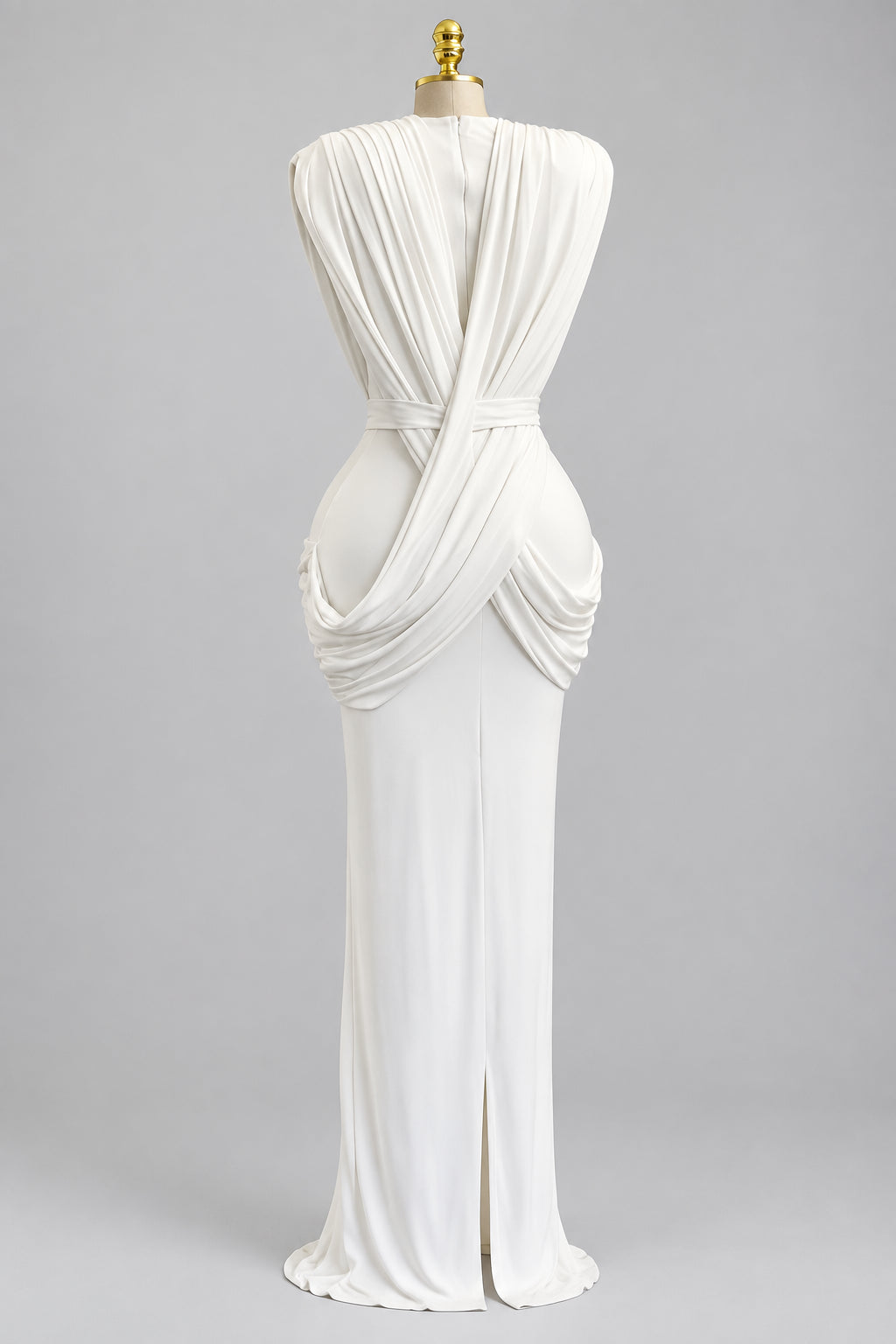 A white draped maxi dress displayed on a mannequin in an indoor setting with beige curtains, a plant, and a small table in the background. The brand name 'CM.