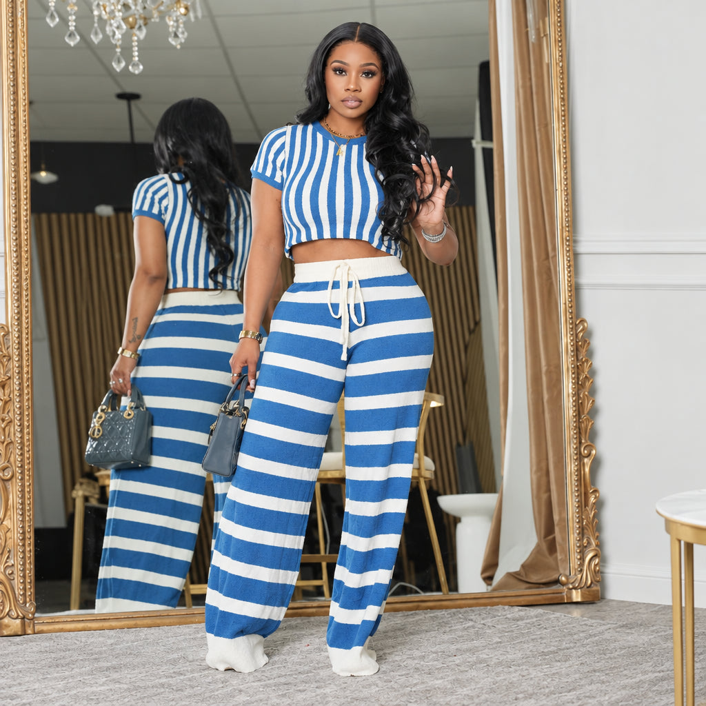 A woman modeling a matching two-piece lounge set with blue and white stripes, consisting of a short-sleeve crop top and wide-leg drawstring pants, standing in front of a mirror in an indoor setting.