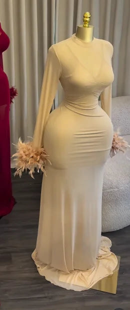 A long-sleeve champagne beige maternity gown displayed on a mannequin, featuring a ruched bodice and matching feathered cuffs, set against a neutral indoor background.
