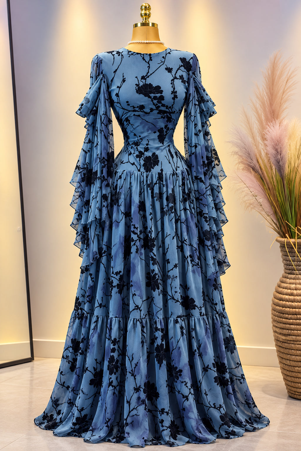 A long-sleeve blue dress with a black floral branch print, displayed on a mannequin. The dress features a fitted bodice, a pleated waist, and sheer flutter sleeves.