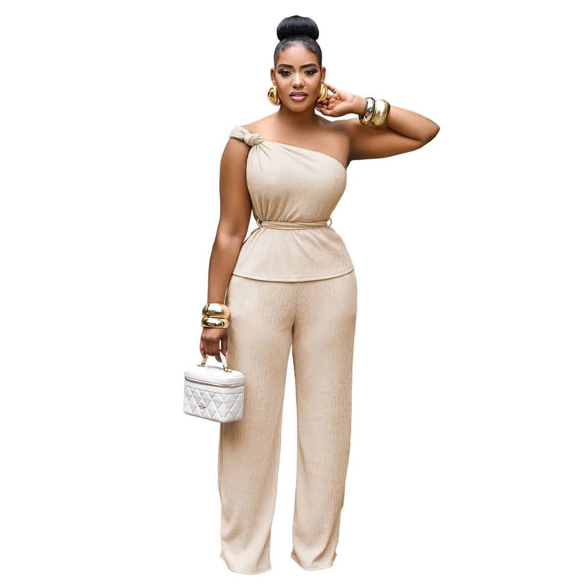 Woman wearing a beige one-shoulder jumpsuit with gold accessories on a white background