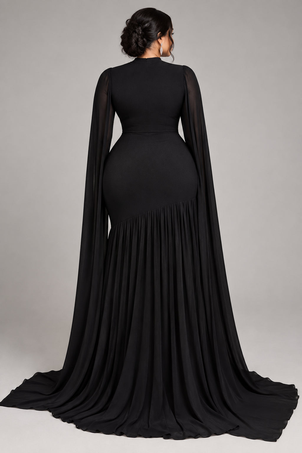 A floor-length black gown displayed on a mannequin in an indoor room with beige curtains, wood flooring, and a potted plant. The dress has a fitted bodice, long draped cape-like sleeves, and a long train.