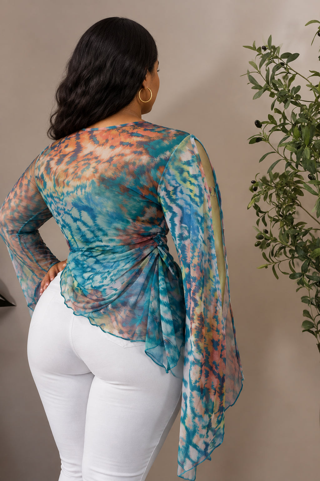 A long-sleeve sheer wrap-style top with a blue, teal, pink, and orange tie-dye pattern, displayed on a gold mannequin torso with white pants. The brand name 'CM.