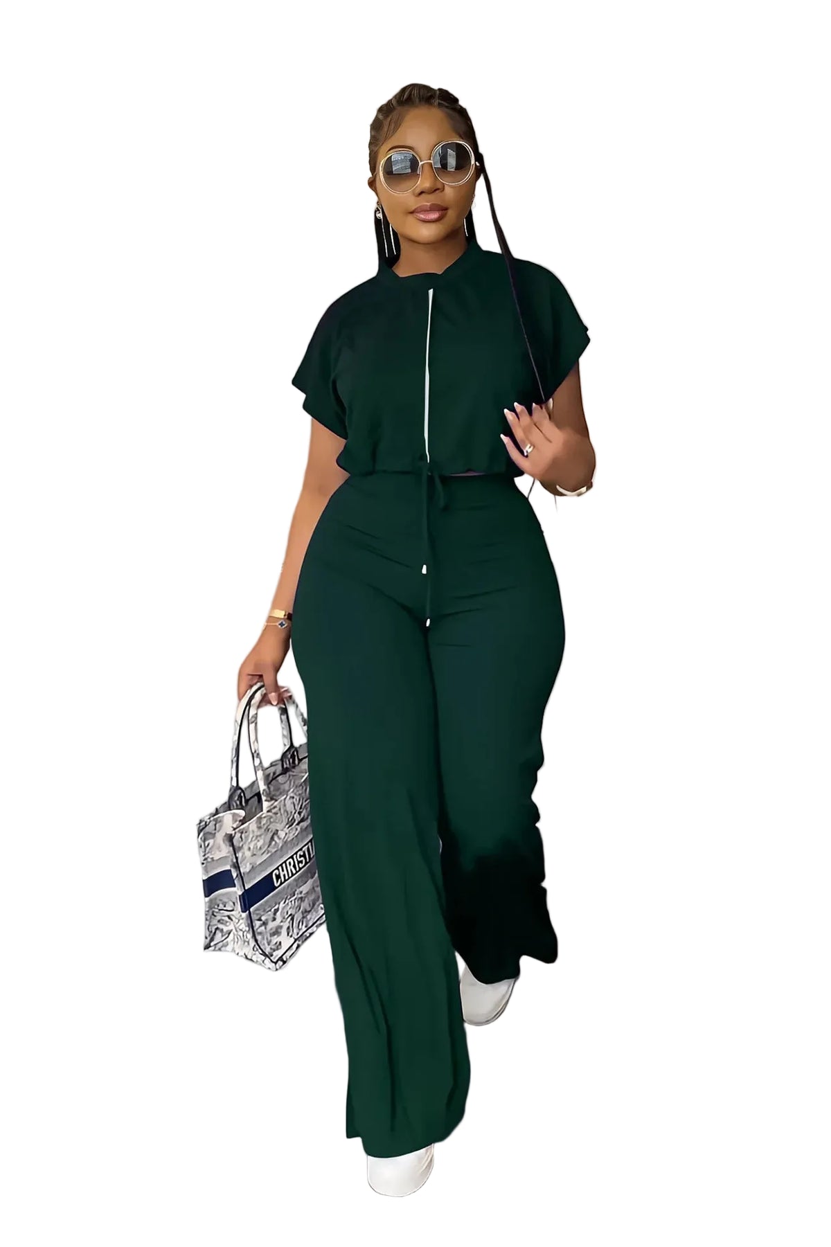 A woman modeling a dark green two-piece jumpsuit with a front zipper and matching high-waisted trousers, styled with sunglasses, a patterned handbag, and white sneakers in an indoor setting.