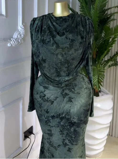 A dark green long-sleeve dress with a tonal floral jacquard pattern, displayed on a mannequin in an indoor setting next to a potted plant.