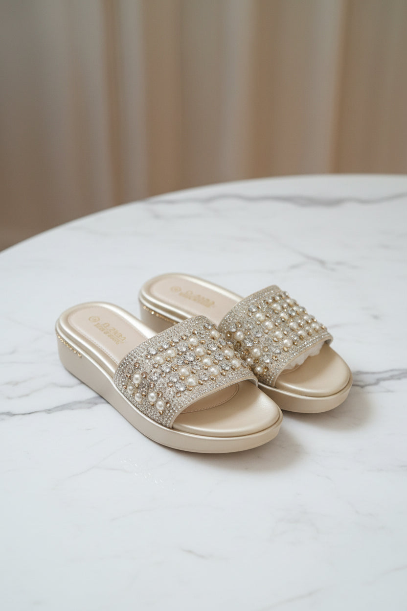 A pair of beige slide sandals with rhinestone embellishments on the straps, still in their clear plastic retail packaging, placed on a dark wood floor.