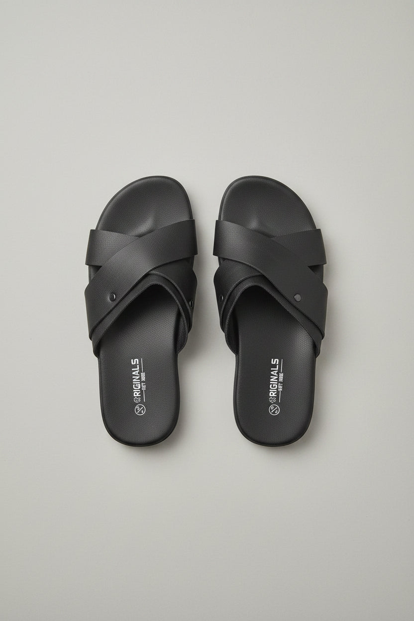 A pair of black slide sandals with wide crisscross straps, placed on a dark wood floor next to a gray upholstered chair.