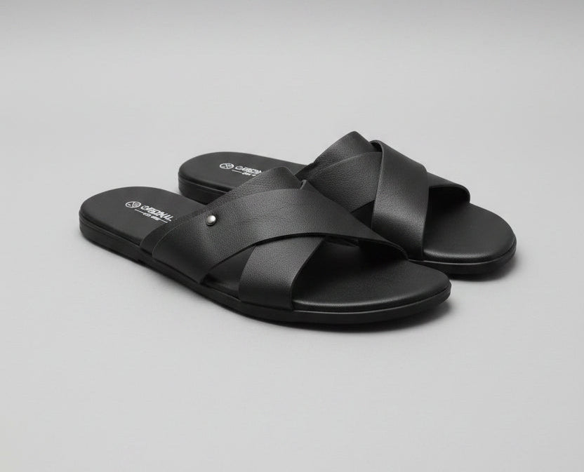 A pair of black slide sandals with crisscross straps, placed on a dark wood floor.