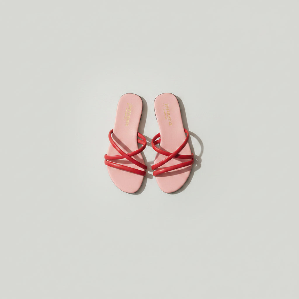 A pair of pink slide sandals with red wrap-around straps, placed on a dark wood floor. The insole features gold branding text that reads 'THE DESIGN' and 'GOLDEN HEART'.