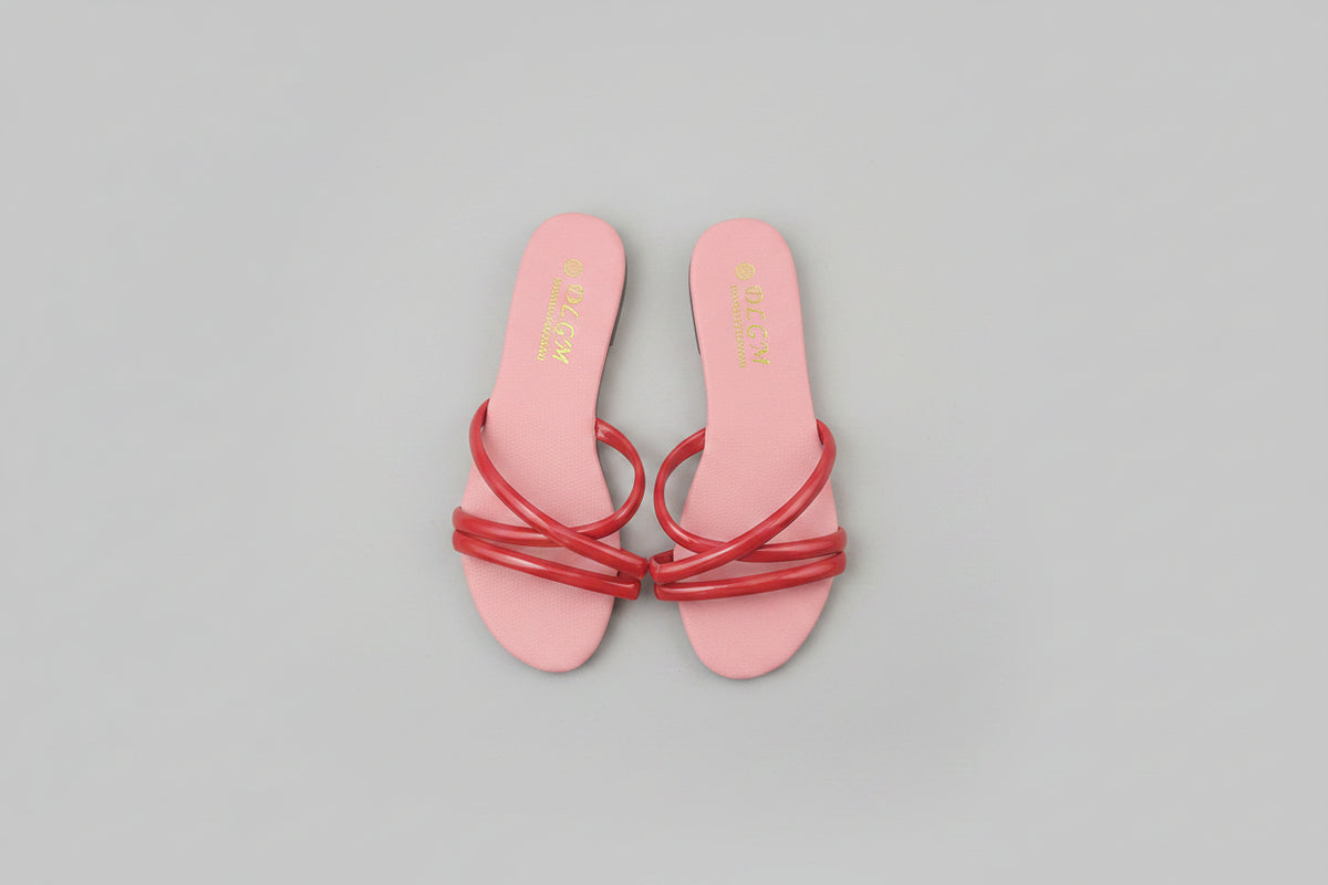 A pair of pink slide sandals with red crisscross straps, placed on a dark wood floor. The brand name 'SOLACE' is printed in gold on the insole.