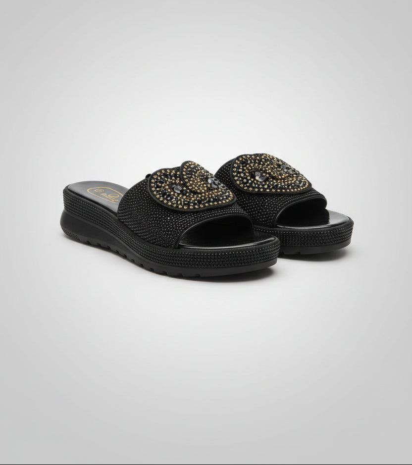 A pair of black women's slide sandals with gold embellishments on the straps, shown from the back on a dark wood floor.