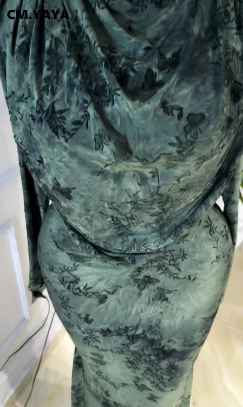 A close-up of a person wearing a long-sleeve, floor-length dress with a cowl neck and an abstract painterly pattern in muted green, gray, and black. The brand name 'CM.