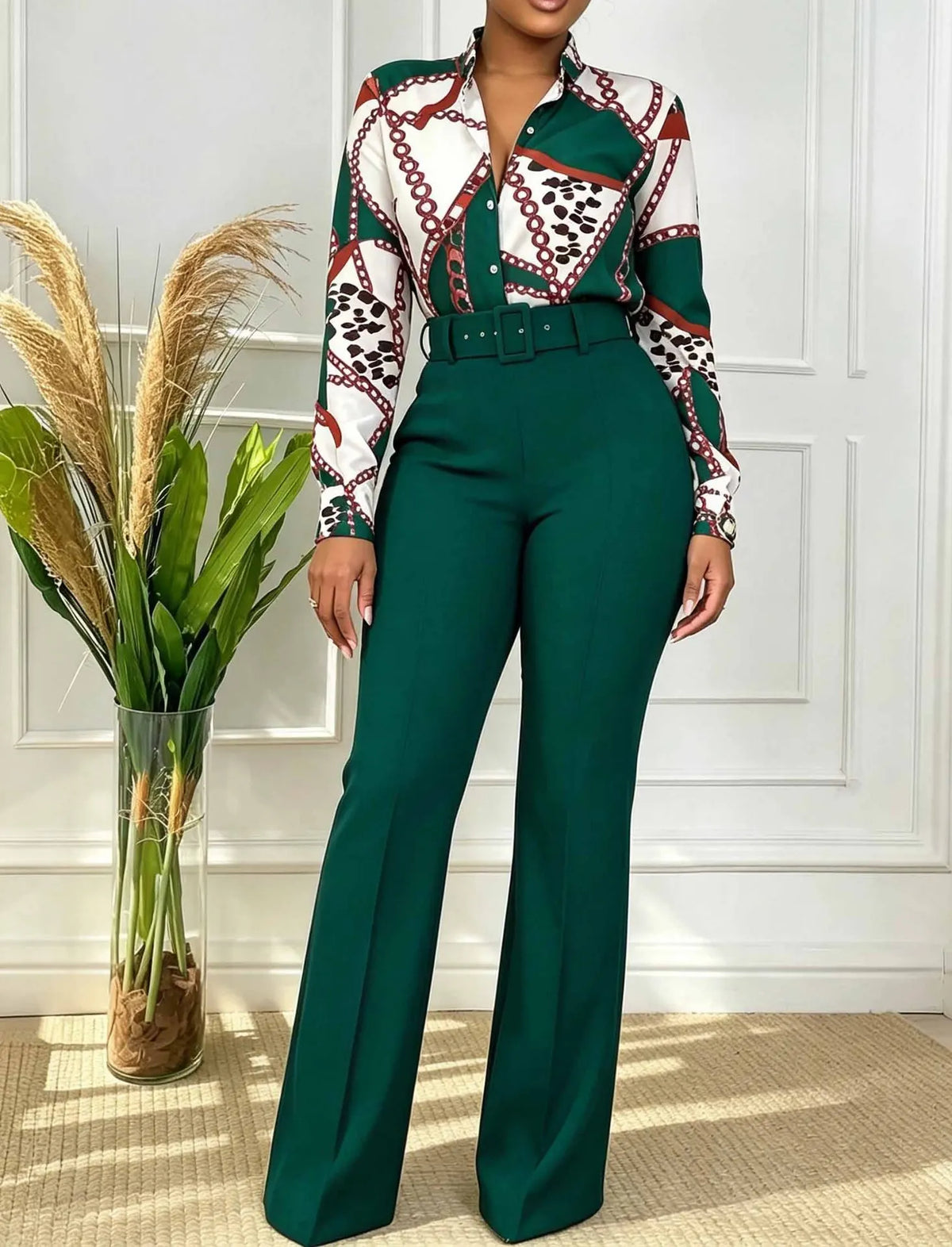 A woman models a matching two-piece outfit consisting of a long-sleeve button-up shirt with a green, white, and red geometric pattern and a matching green belt, paired with high-waisted green wide-leg trousers. She stands in front of a white paneled wall next to a glass vase with dried pampas grass.