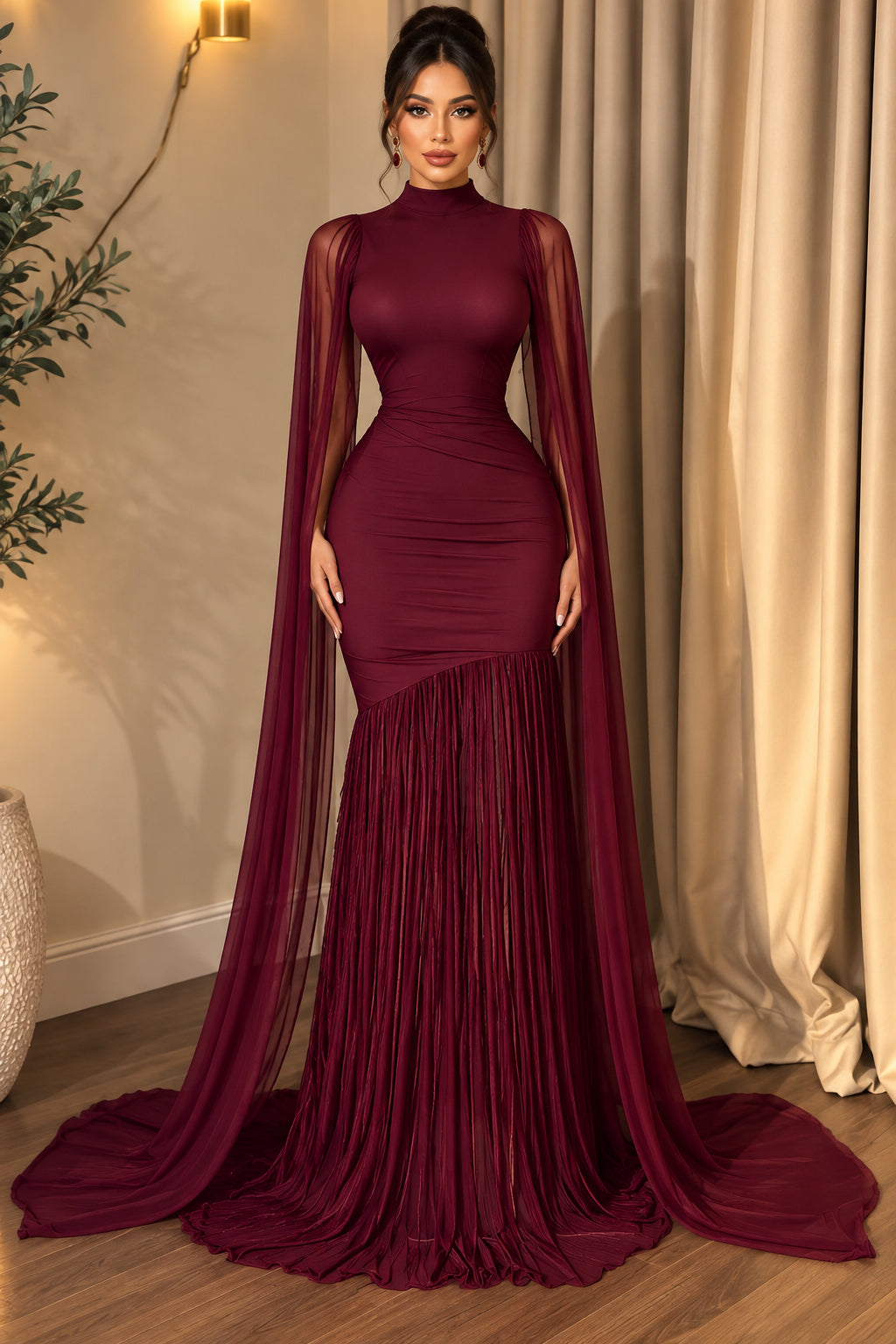 A deep burgundy evening gown displayed on a mannequin in an indoor setting with beige curtains and a wooden floor. The dress features a high neckline, a fitted waist, a long sheer draped capelet, and a pleated mermaid-style skirt with a long train.