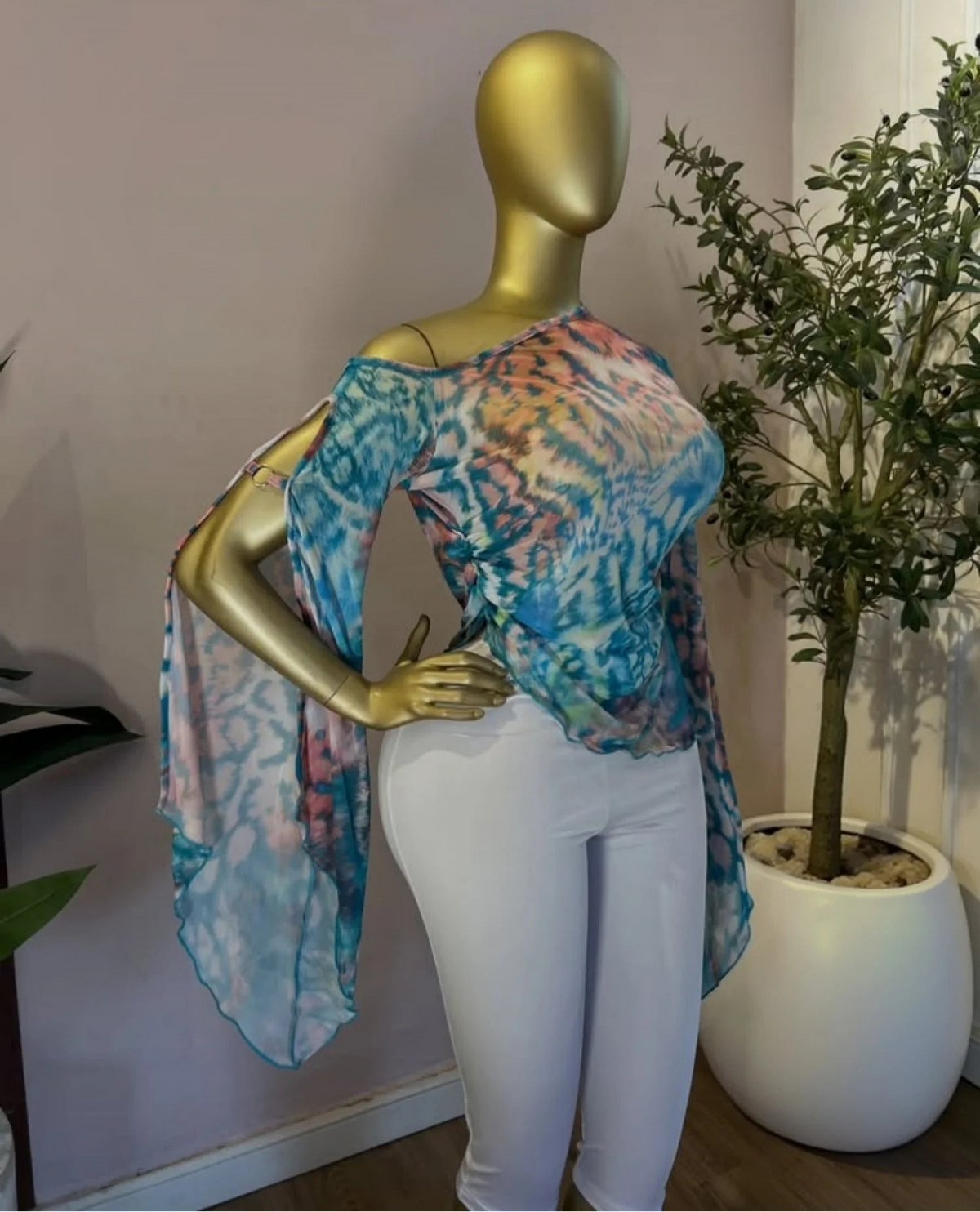 A gold mannequin wearing a sheer, off-the-shoulder blue and pink tie-dye top with white pants, standing indoors next to a potted plant. The brand name 'CM.