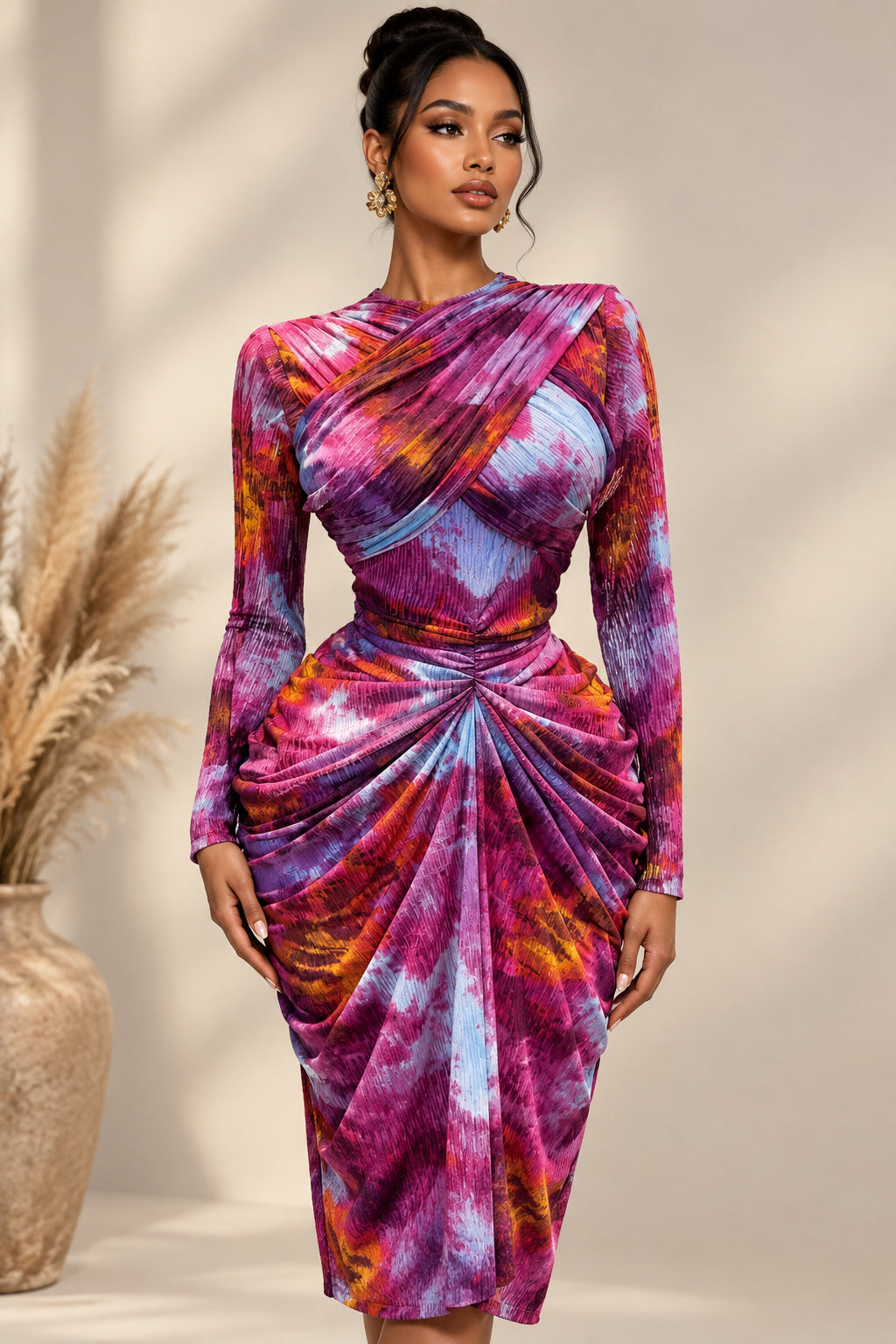 A long-sleeve draped dress displayed on a mannequin, featuring a vibrant purple, pink, orange, and blue tie-dye pattern. The dress has a wrap-style bodice with long sleeves and a gathered skirt, set against a background with decorative pampas grass in woven vases.