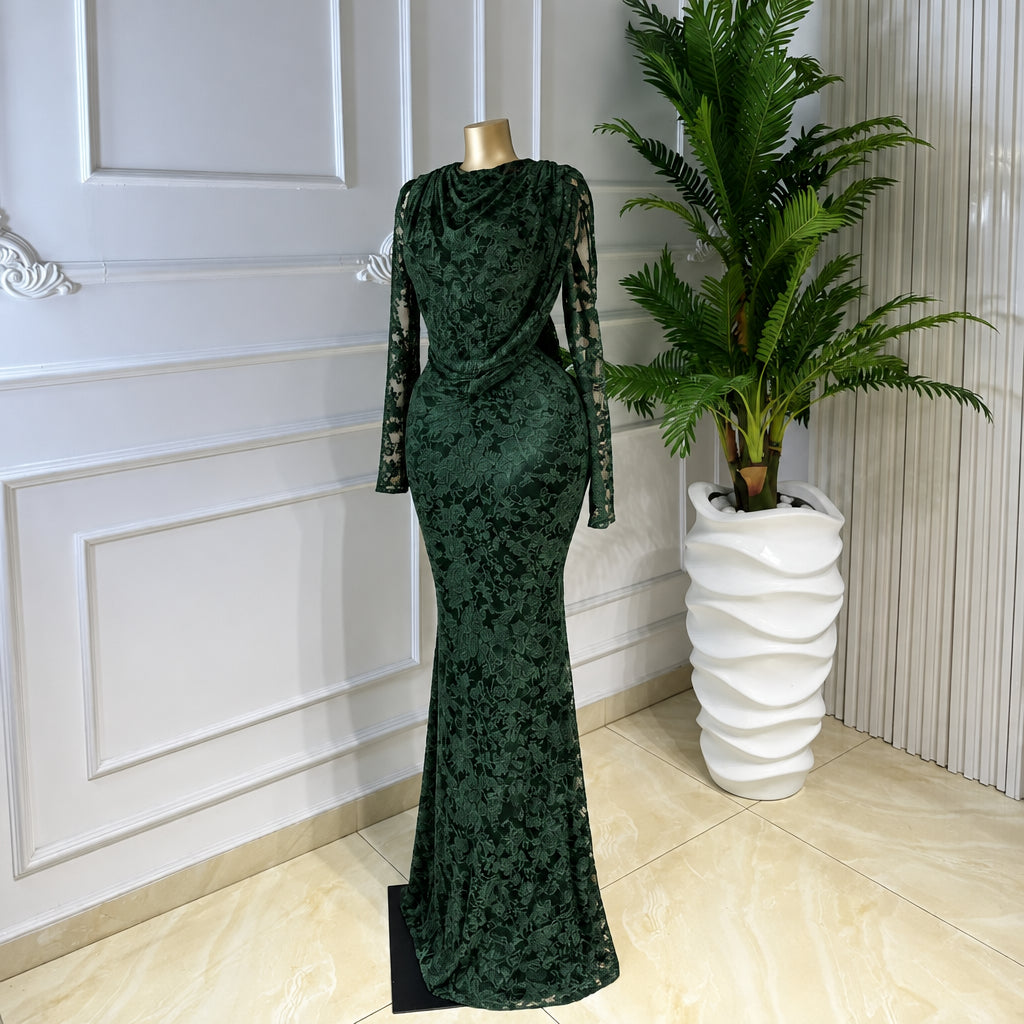 A long-sleeve floor-length gown in a dark green marbled velvet fabric, displayed on a mannequin in an indoor setting with a white paneled wall, tiled floor, and a potted plant.