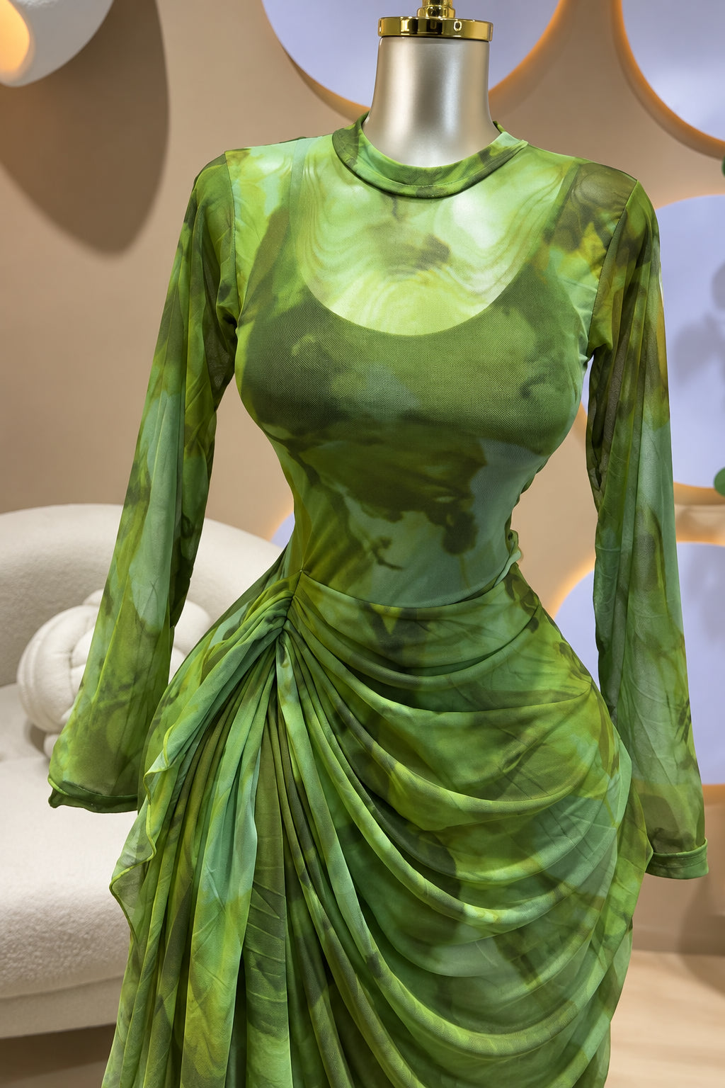 A long-sleeve green dress with an abstract watercolor-like print in shades of olive, moss, and lime, displayed on a metallic mannequin torso. The dress features a scoop neckline, fitted bodice, and a draped, gathered skirt.