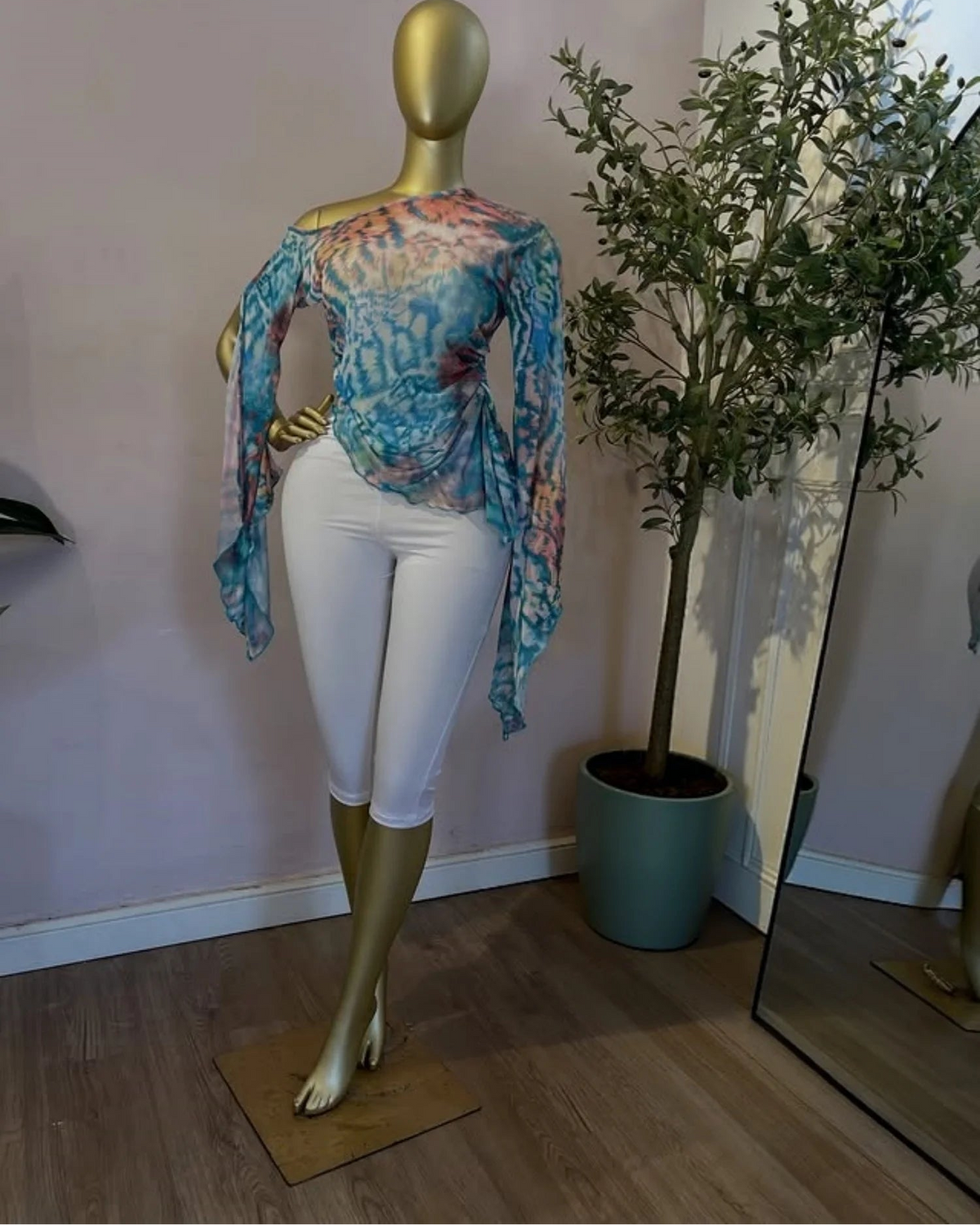 A gold mannequin wearing a colorful tie-dye long-sleeve top and white capri leggings stands indoors next to a potted tree. The brand name 'CM.