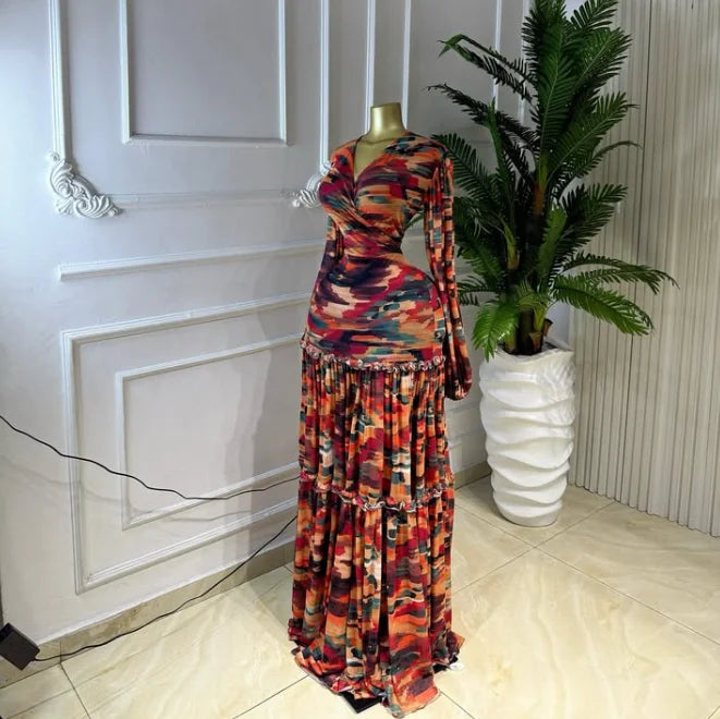 A long-sleeve maxi dress with a colorful abstract camouflage pattern in red, orange, purple, and black, displayed on a mannequin in an indoor setting next to a potted plant.