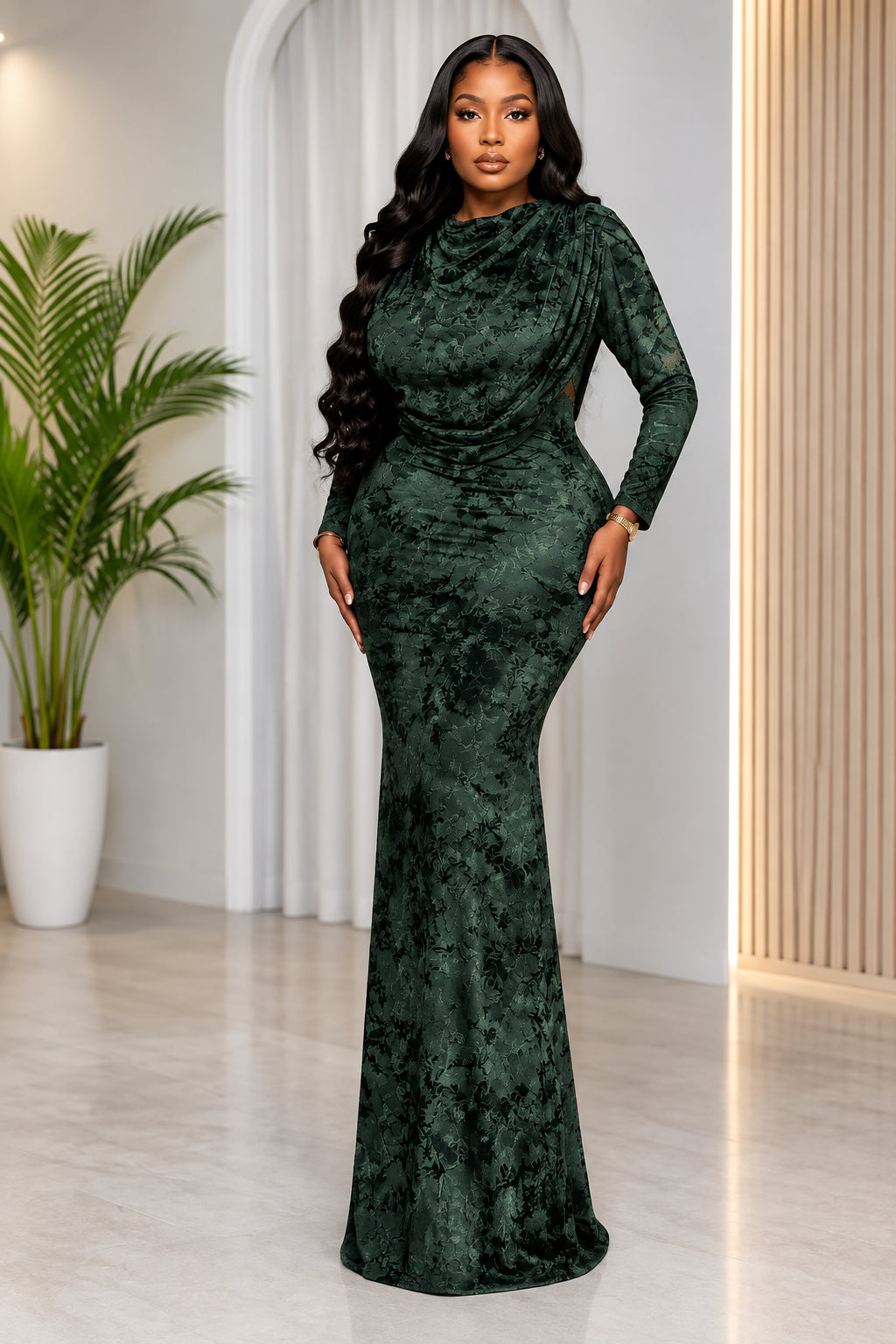 A woman modeling a dark green crushed velvet long-sleeve mermaid gown in an indoor setting with a potted plant and neutral walls. The brand name 'CM.