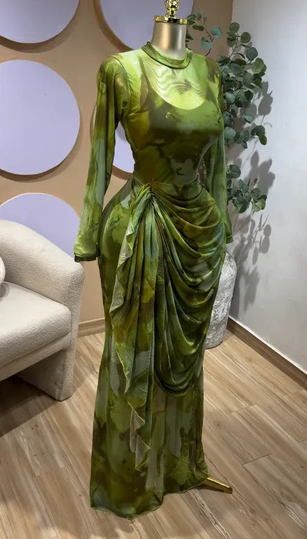 A long-sleeved floor-length dress in a green and olive abstract print, displayed on a mannequin in an indoor showroom setting with a beige armchair, wood flooring, and decorative wall panels.