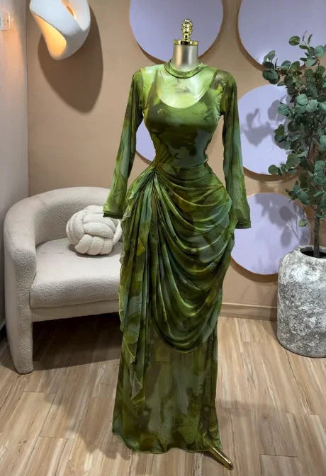 A floor-length green satin gown with long sleeves and a draped waist detail, displayed on a mannequin in an indoor showroom setting with a chair, wall decor, and plant.