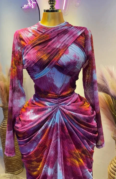 A long-sleeve draped dress displayed on a mannequin, featuring a vibrant purple, pink, orange, and blue tie-dye pattern.