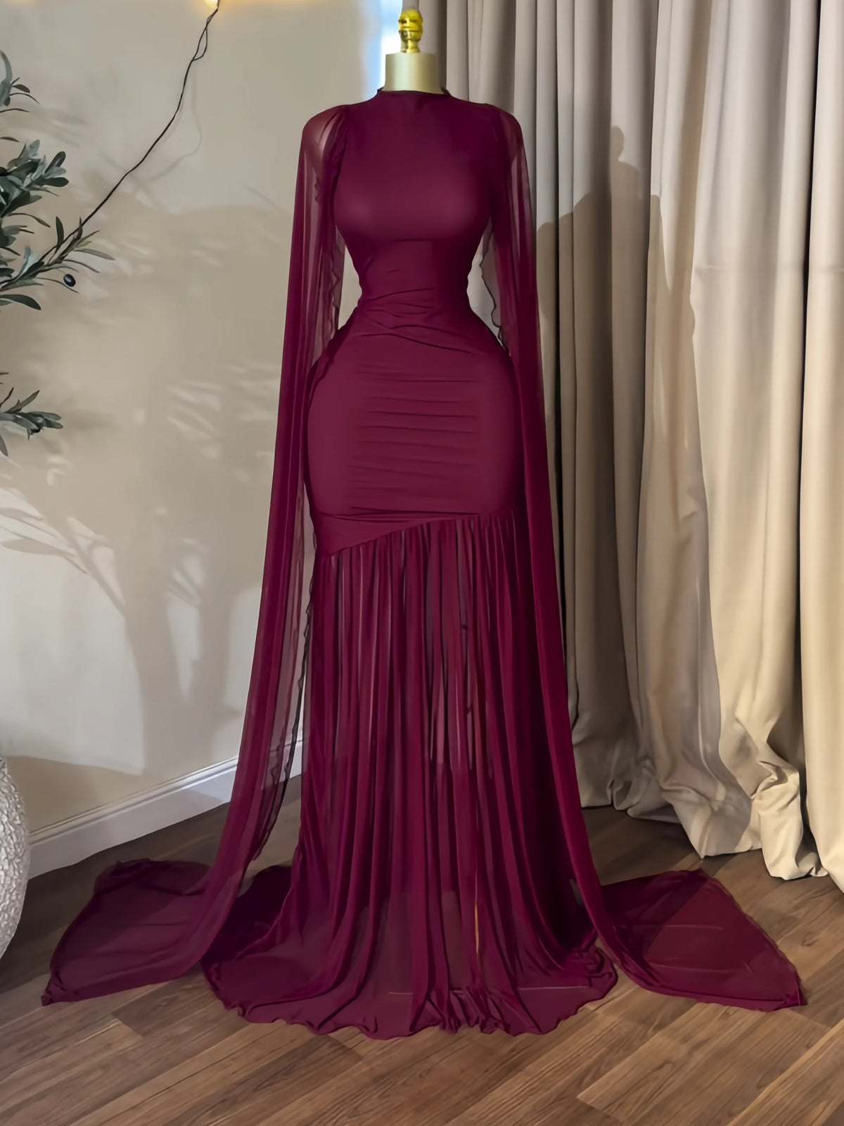A floor-length burgundy gown displayed on a mannequin indoors, featuring a fitted bodice, long sheer draped sleeves, a pleated mermaid-style skirt, and a long train on a wood floor with beige curtains behind.