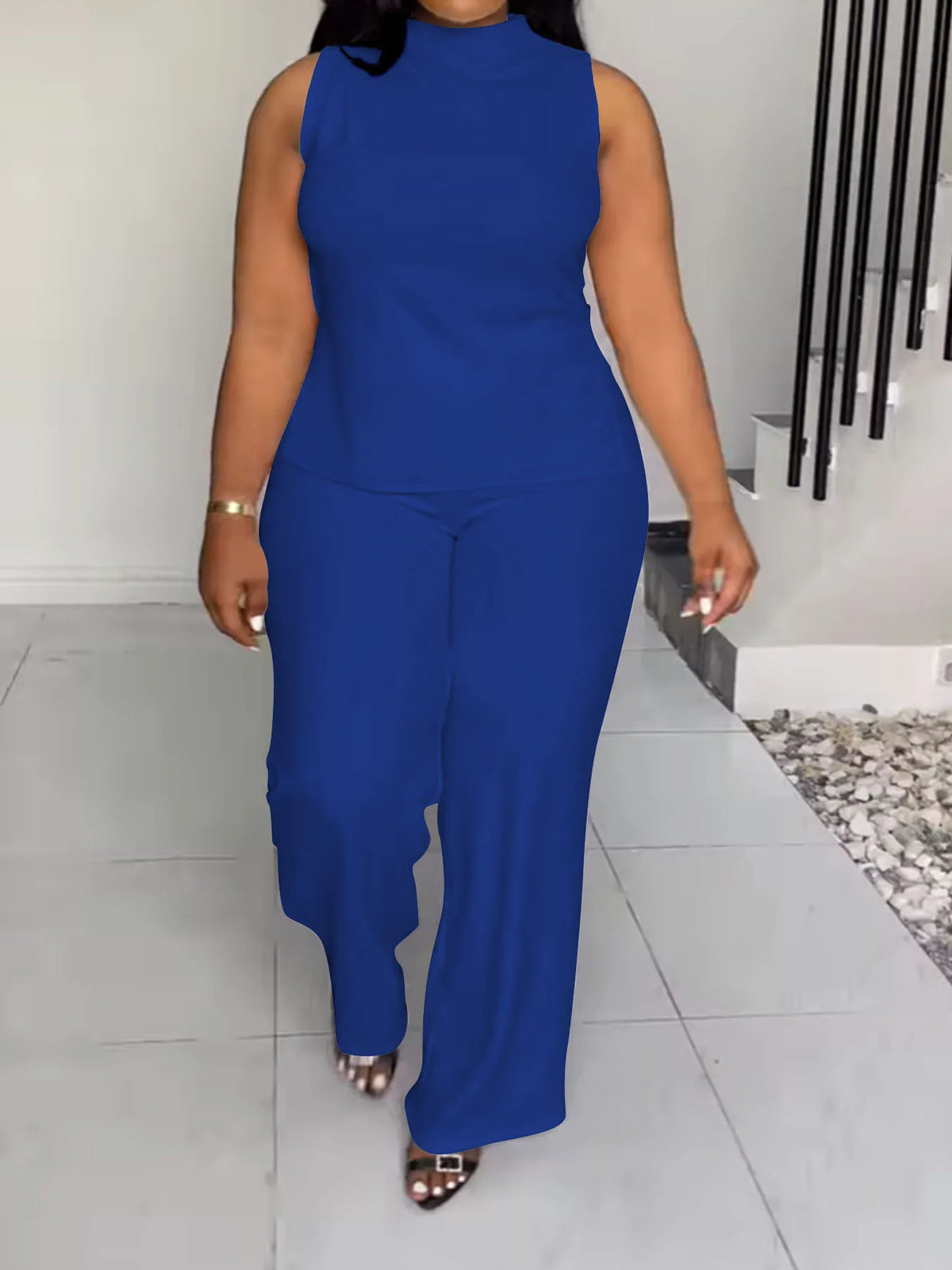 A woman modeling a sleeveless royal blue jumpsuit with a mock neck and wide-leg pants, walking in a modern indoor setting with light gray tiled floors and a staircase.