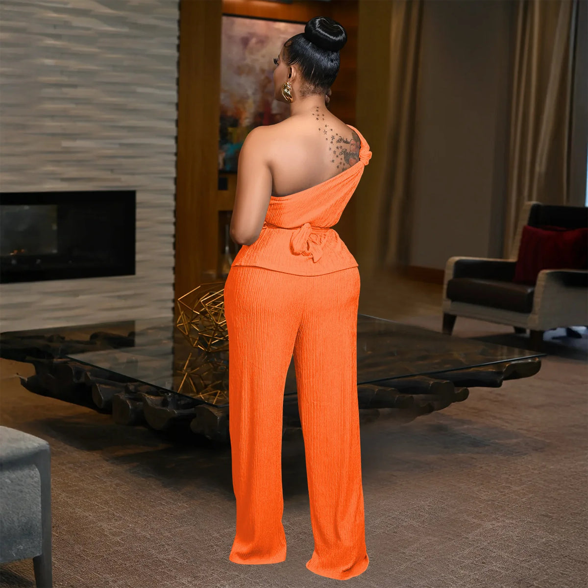 A woman stands with her back to the camera wearing a bright orange one-shoulder jumpsuit in a modern living room setting.