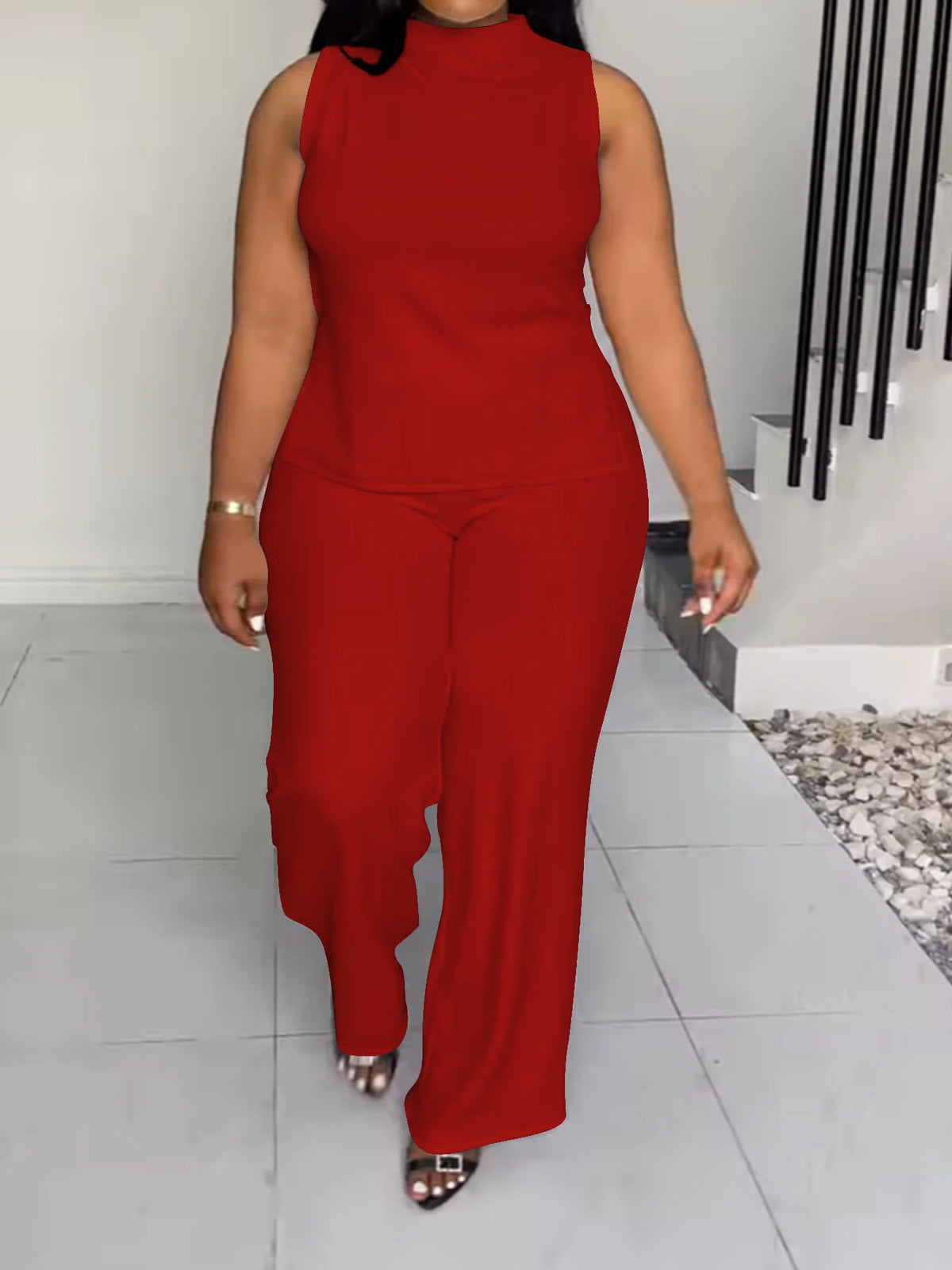 A woman modeling a bright red sleeveless mock-neck jumpsuit with wide-leg pants, walking in a modern indoor setting with light gray tiled floors and a white wall.