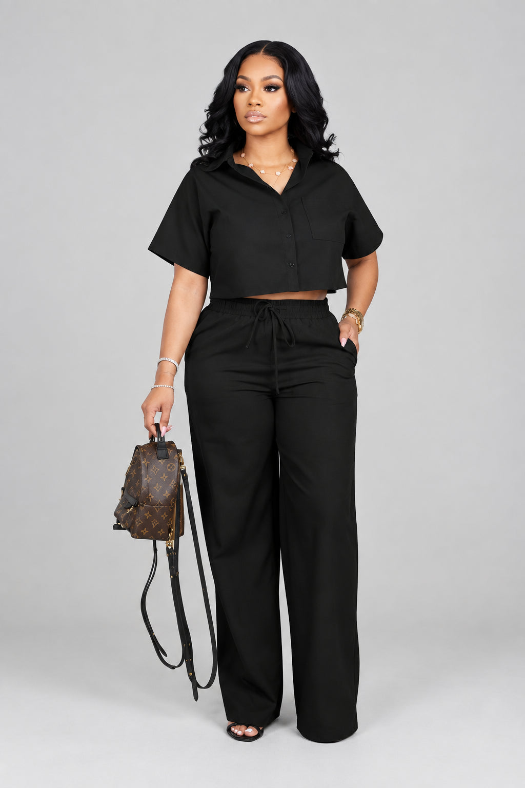 A woman modeling a black two-piece outfit consisting of a short-sleeve collared crop top and matching wide-leg drawstring pants, standing in a modern interior with a slatted wood wall, pink flowers, and a mirror.