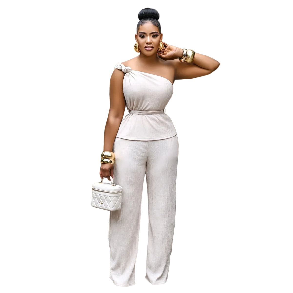 A woman models a light beige ribbed one-shoulder jumpsuit in a modern living room setting. The jumpsuit features a draped off-the-shoulder neckline, a defined waist, a peplum-style midsection, and wide-leg trousers.