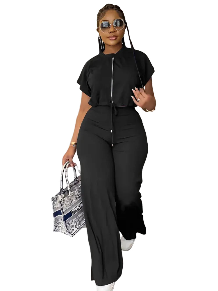 A woman modeling a black short-sleeve jumpsuit with a front zipper and drawstring waist, styled with sunglasses, a patterned tote bag, and white sneakers in an indoor setting.