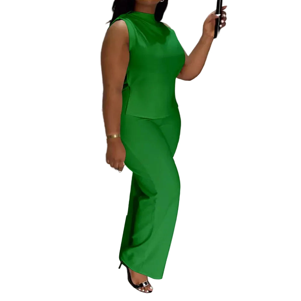 A woman wearing a bright green sleeveless jumpsuit with a high mock neck and wide-leg pants, standing indoors near a staircase with black railings. The brand name 'CM.