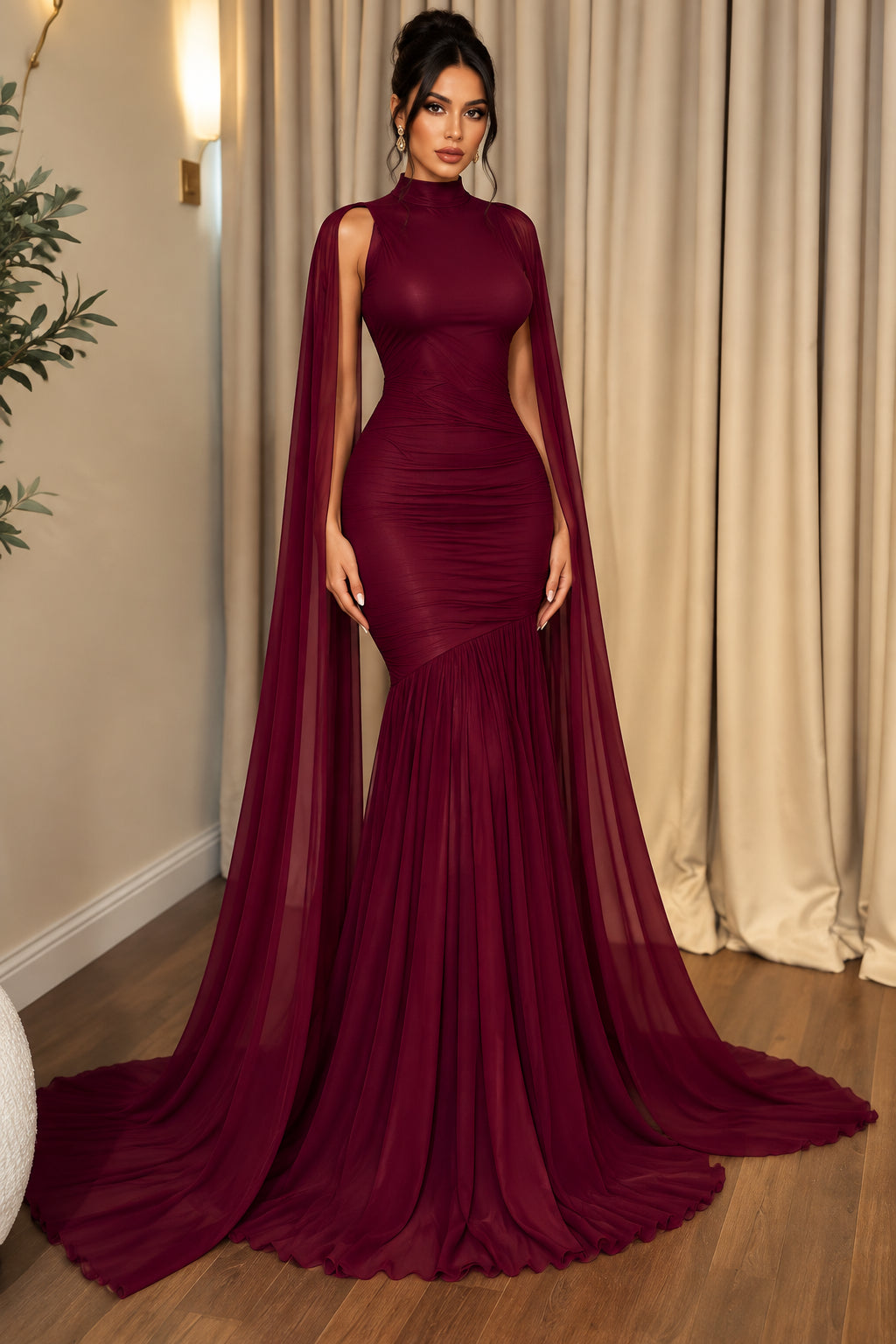 A floor-length magenta gown displayed on a mannequin in an indoor room with beige curtains and wood flooring. The dress has long sheer draped sleeves, a fitted bodice, and a long train.