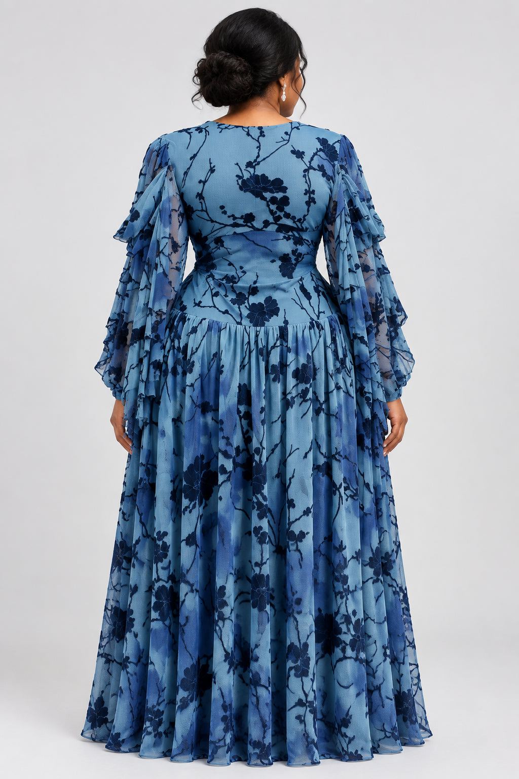 A blue long-sleeve maxi dress with a black floral branch pattern, displayed on a mannequin with a pearl necklace. The brand name 'CM.