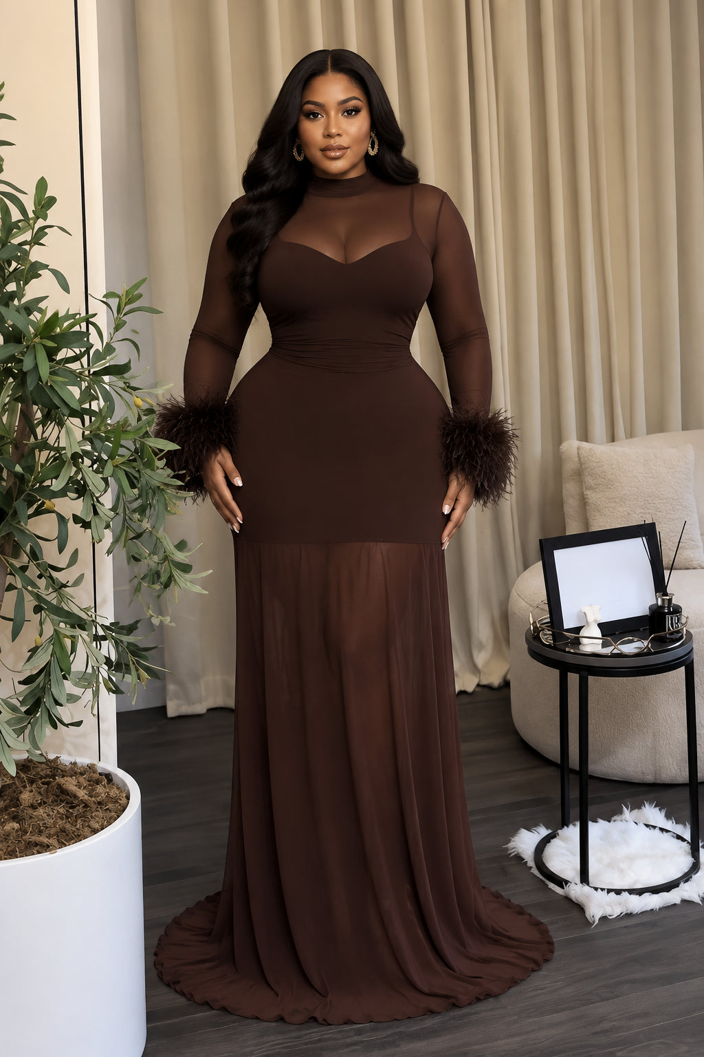 A floor-length dark brown evening gown displayed on a mannequin in an indoor room setting. The dress has long sheer sleeves, a fitted bodice, feathered cuffs, and a flowing skirt with a slight train.