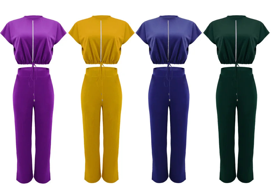 Five color options of a two-piece scrub set by CM.YAYA, featuring a short-sleeve crop top and matching pants in black, purple, mustard yellow, royal blue, and forest green, shown on a plain white background.