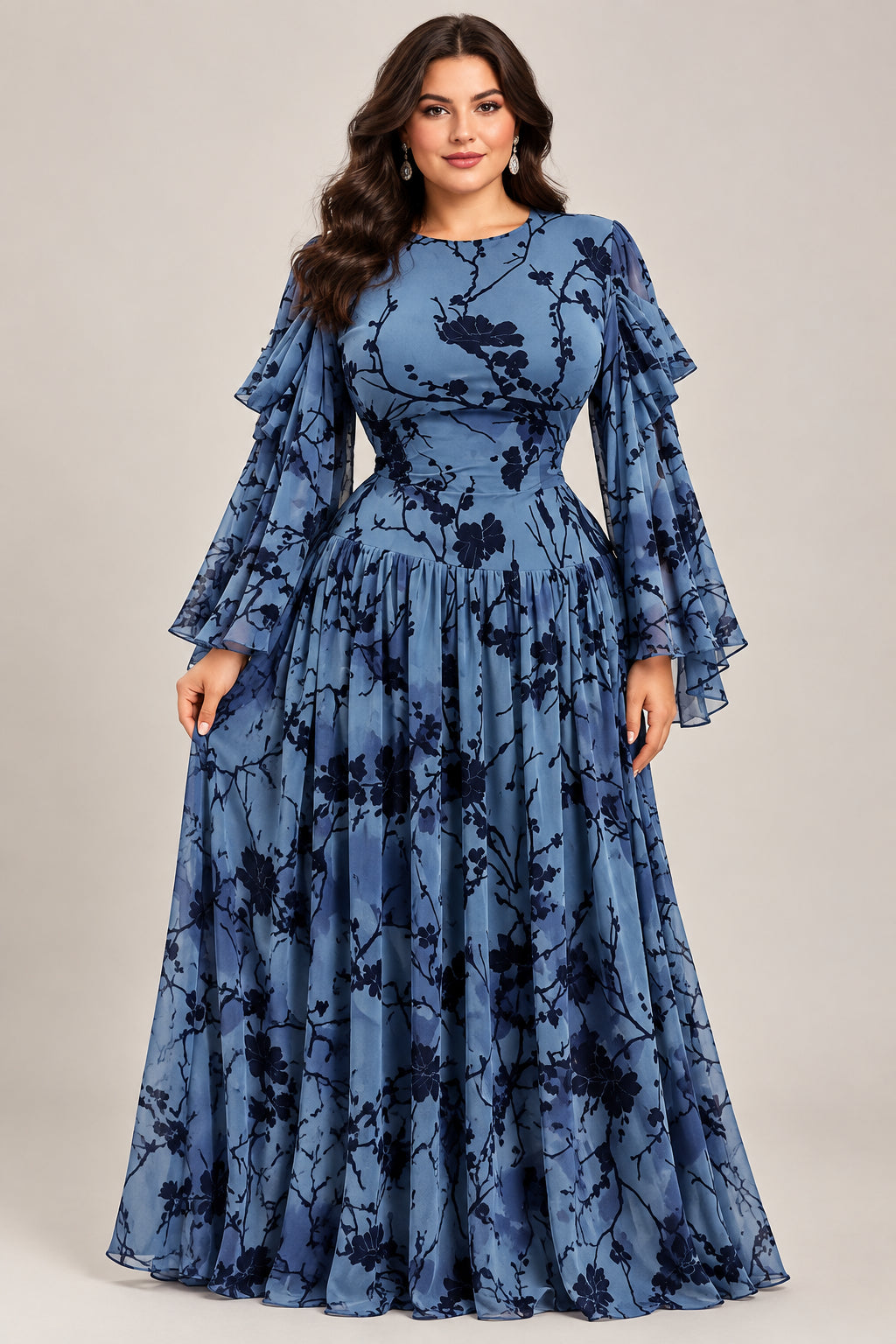 A long-sleeve blue maxi dress with a black floral branch print is displayed on a mannequin in an indoor boutique setting. The dress features a fitted bodice, ruffled sleeves, and a flowing skirt.