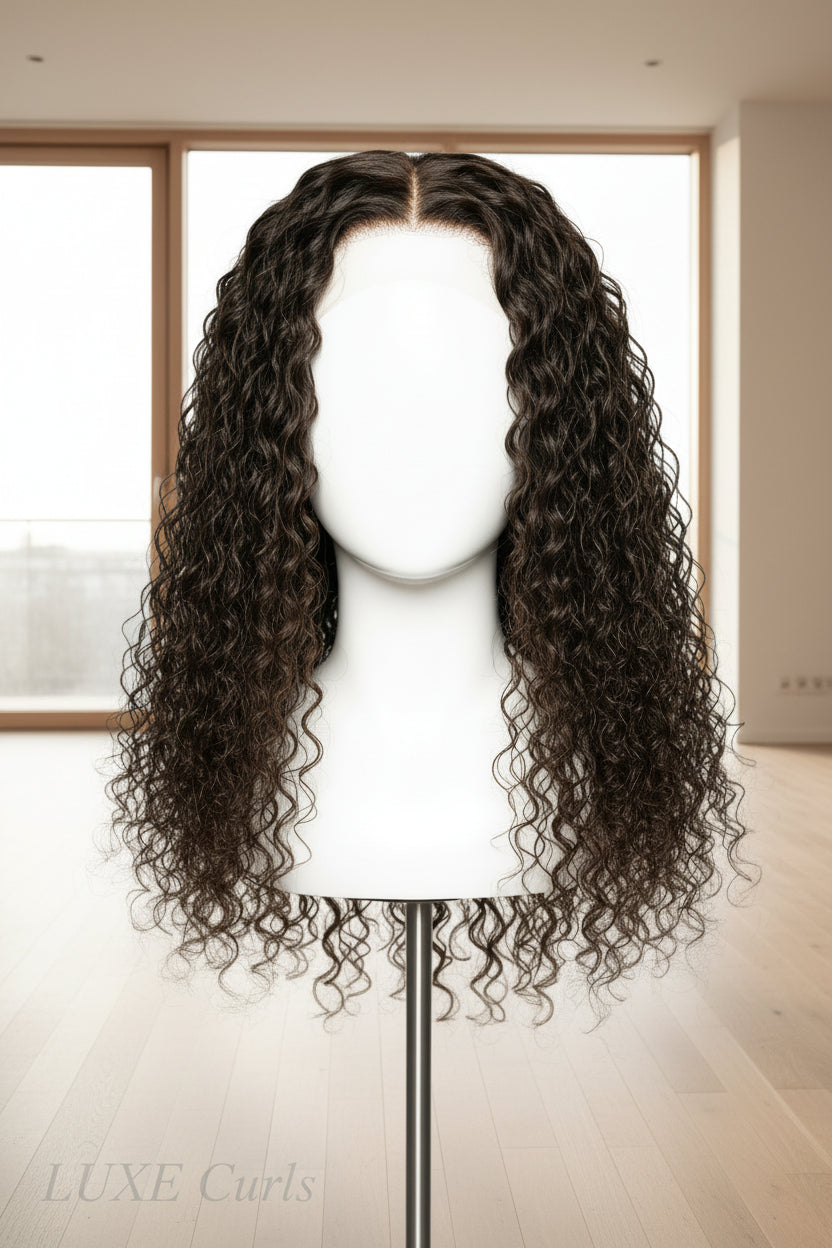 A long, dark brown to black curly wig displayed on a stand against a plain light gray wall.