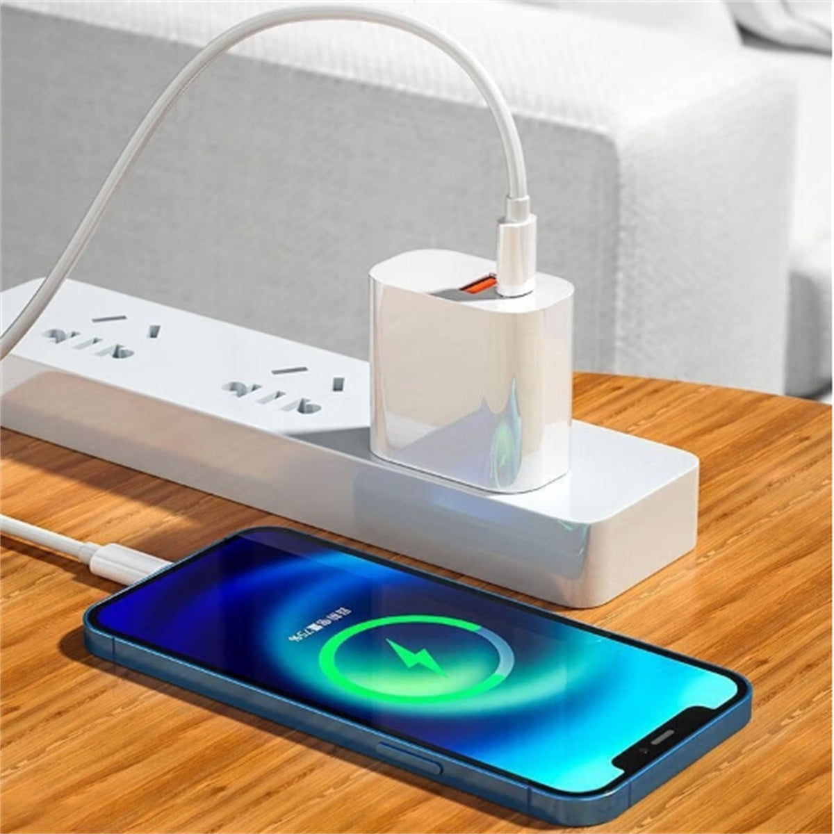 A white multi-port power adapter sits on a wooden table, plugged into a wall outlet and connected to a smartphone showing a wireless charging screen. A white charging cable loops up toward a sofa in the background.